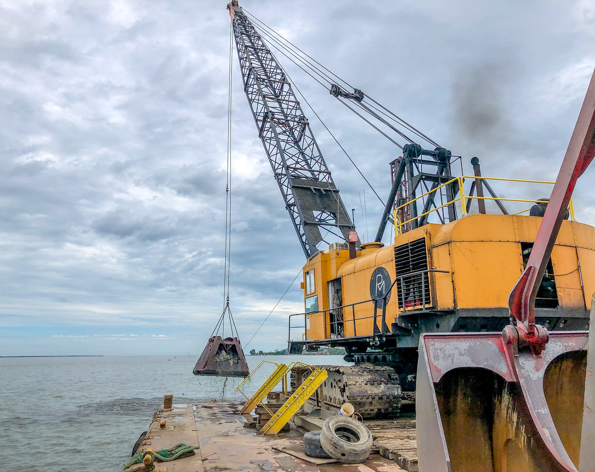 A yellow crane lifts a clamshell bucket beside a dredging barge in Sandusky Harbor.